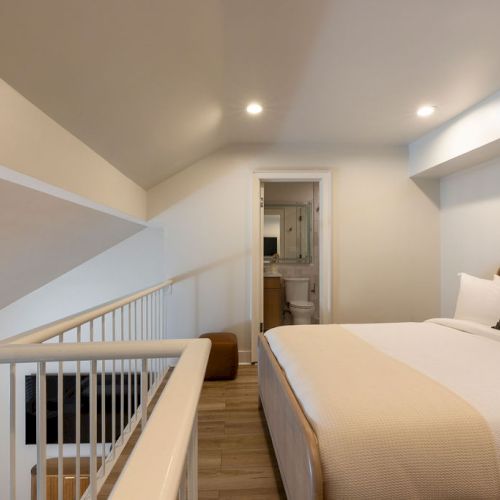 The image shows a cozy loft bedroom with a bed, framed art, and a small hallway leading to a bathroom.