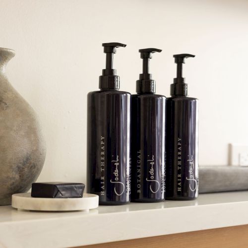 The image shows three black pump bottles labeled "Hair Therapy" and a dark bar on a tray, placed on a shelf next to a vase.