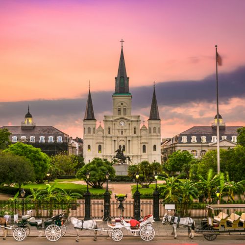 A historic cathedral stands tall with surrounding lush gardens at sunset, and horse-drawn carriages line the street in front.