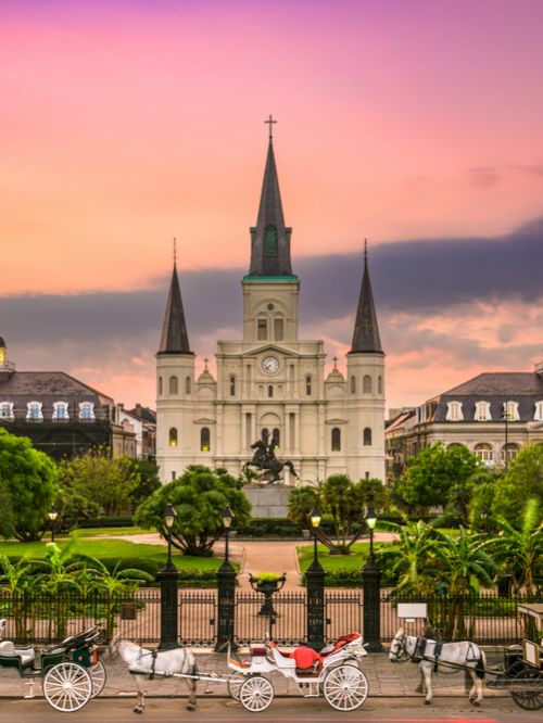 A historic cathedral stands tall with surrounding lush gardens at sunset, and horse-drawn carriages line the street in front.