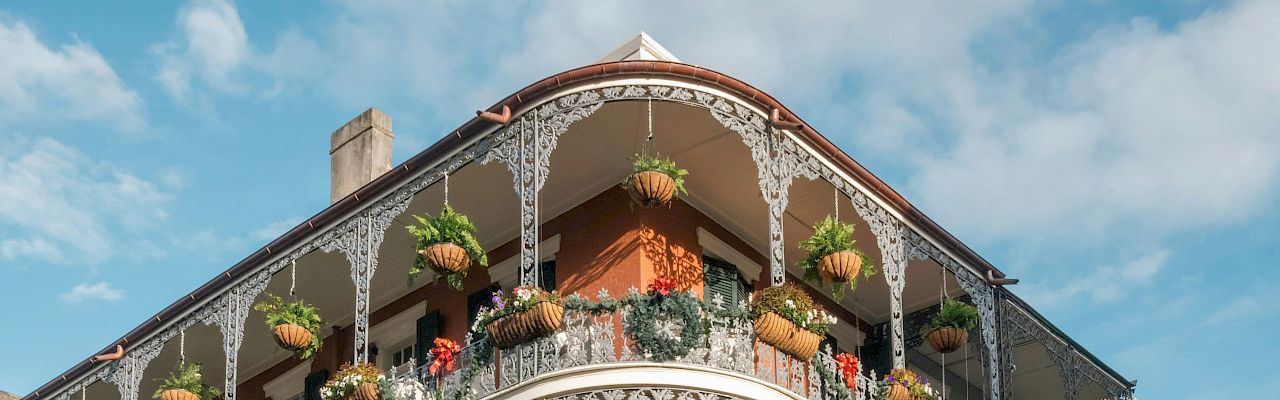 A historic building with ornate iron balconies adorned with hanging plants and flowers, set against a cloudy blue sky backdrop.