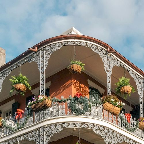 A historic building with ornate iron balconies adorned with hanging plants and flowers, set against a cloudy blue sky backdrop.