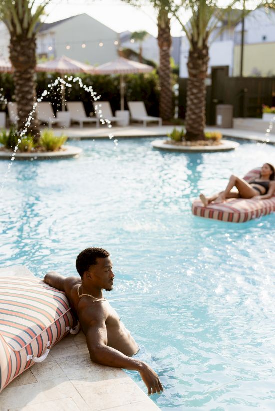 A person relaxes on a pool's edge while another enjoys a float on the water, surrounded by loungers and palm trees in the background.