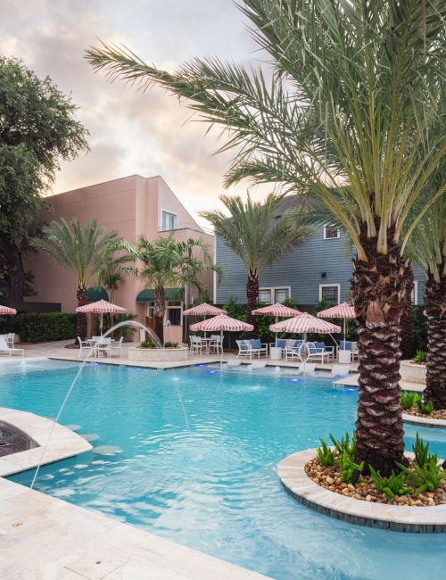 A serene poolside scene with palm trees, lounge chairs, and umbrellas, adjacent to pastel buildings under a partly cloudy sky.