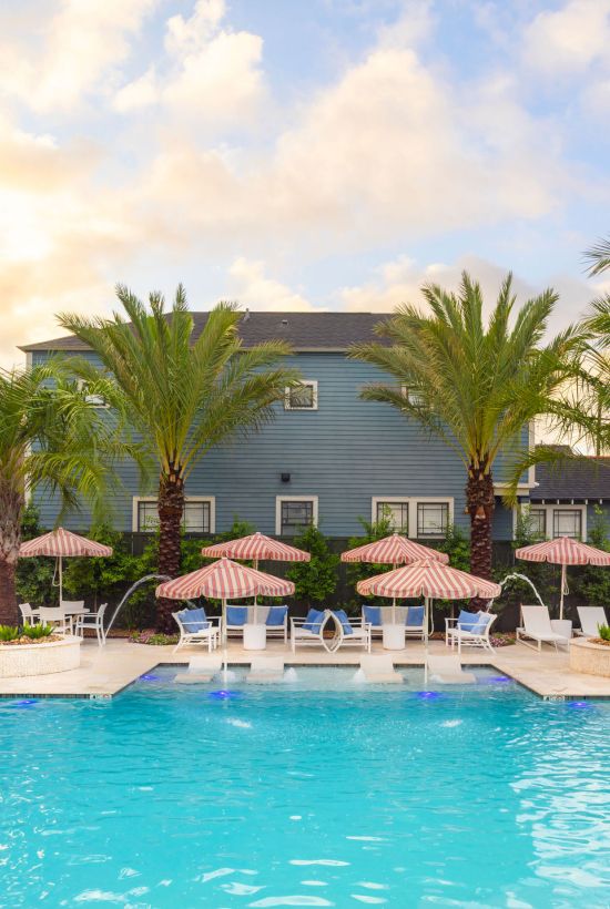 A swimming pool with palm trees and pink-striped umbrellas in front of a blue building is shown in the image.