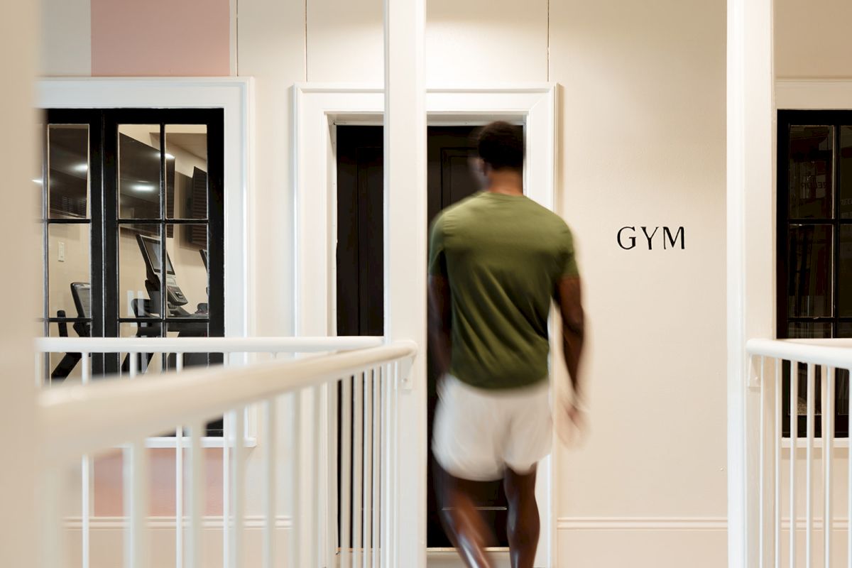 A person in a green shirt and white shorts walks toward a door labeled "GYM" with exercise equipment visible through nearby windows.