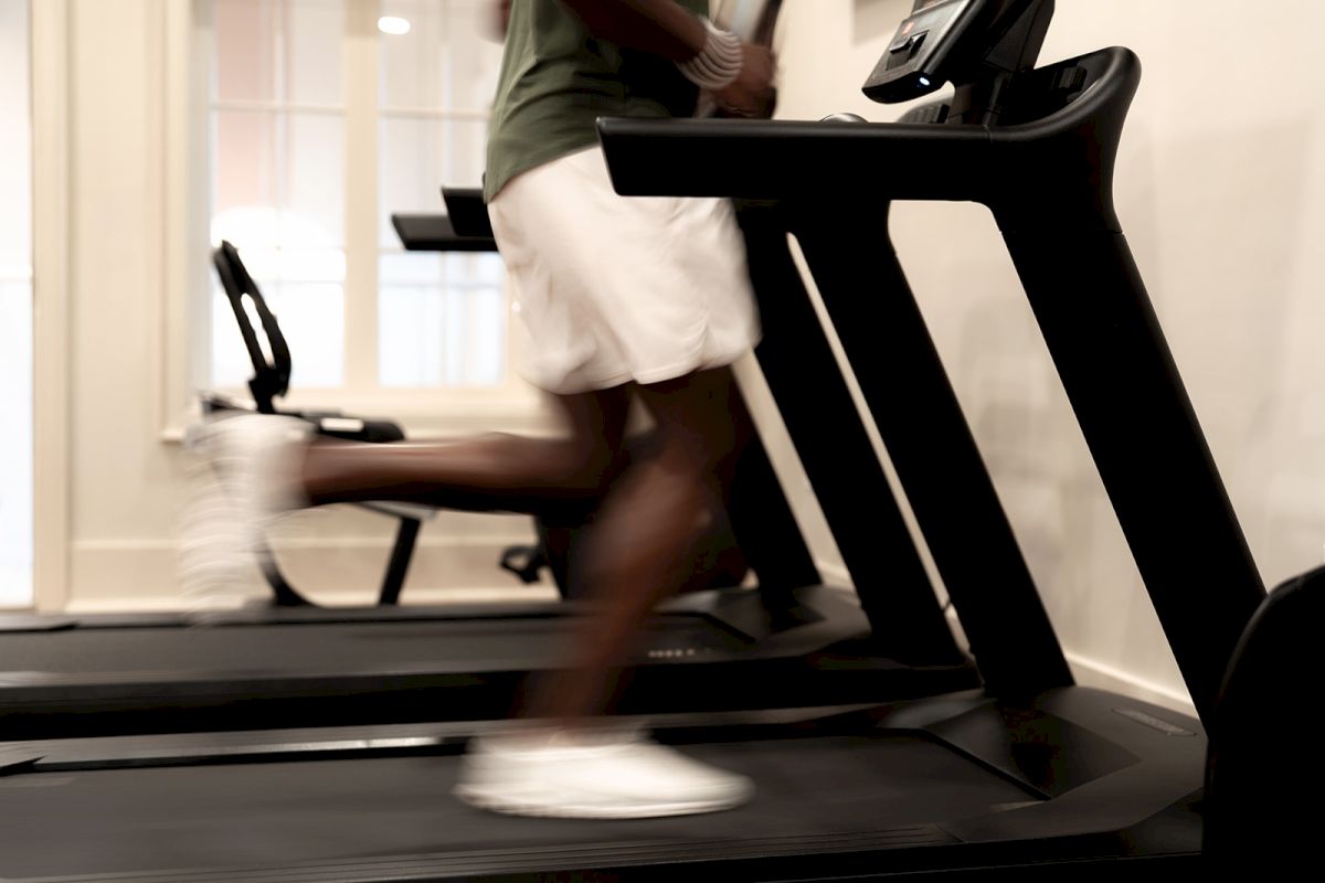 A person is running on a treadmill in a gym setting, wearing a green shirt and white shorts. A stationary bike is visible in the background.