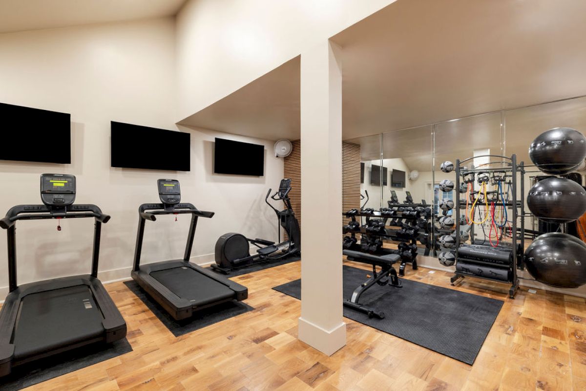 A home gym with treadmills, exercise bike, weights, exercise balls, and mirrors on a wooden floor.