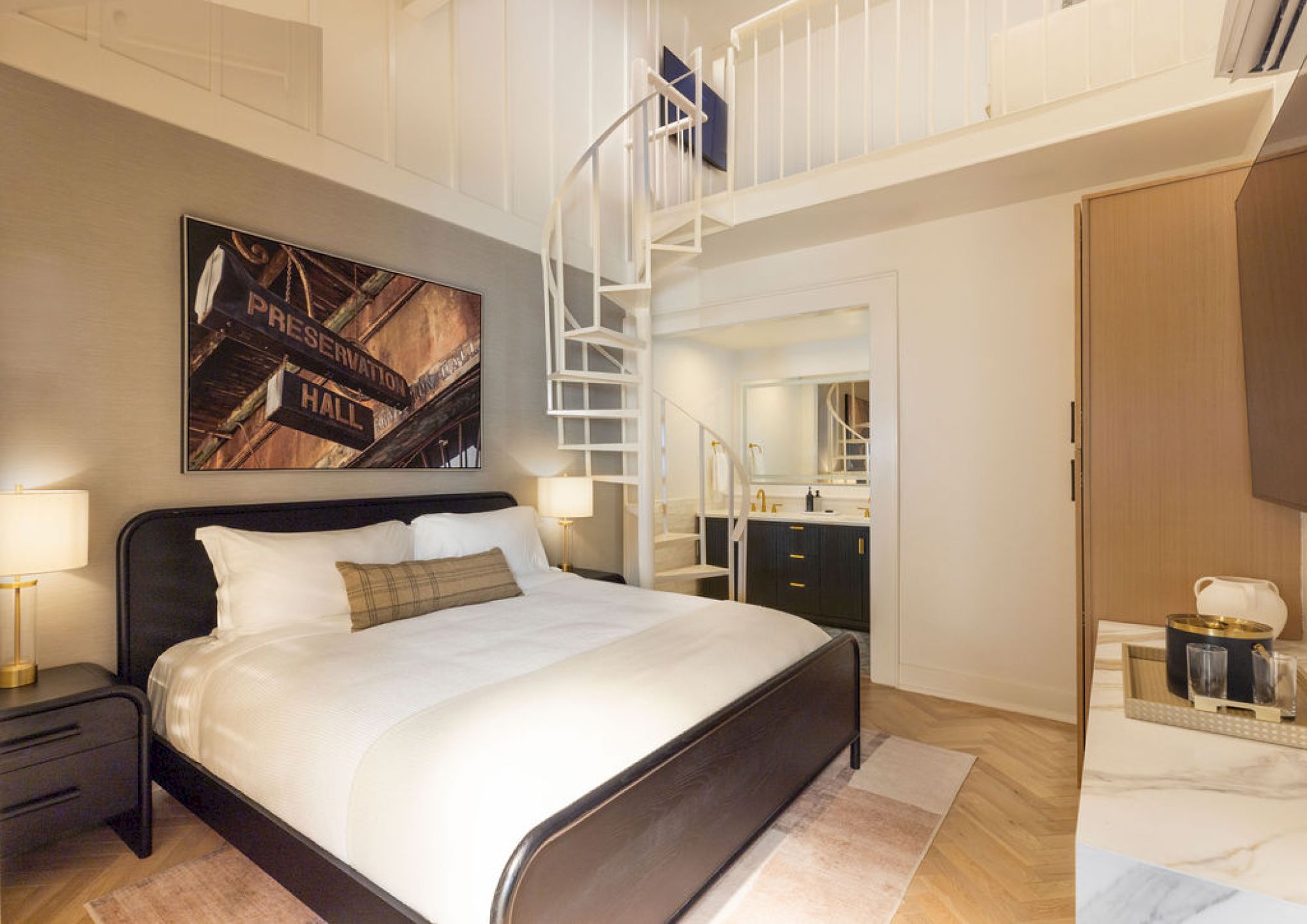 A modern bedroom features a loft, spiral staircase, cozy bed, bedside lamps, artwork on the wall, and an open bathroom area.