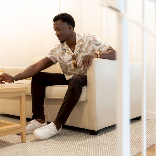 A person sitting on a couch, reaching for a drink on a table, with a bottle beside it, in a bright, minimalist room.