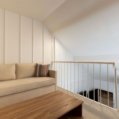 The image shows a cozy loft space with a beige sofa, wooden coffee table, and white railing, featuring a sloped ceiling and minimalist decor.