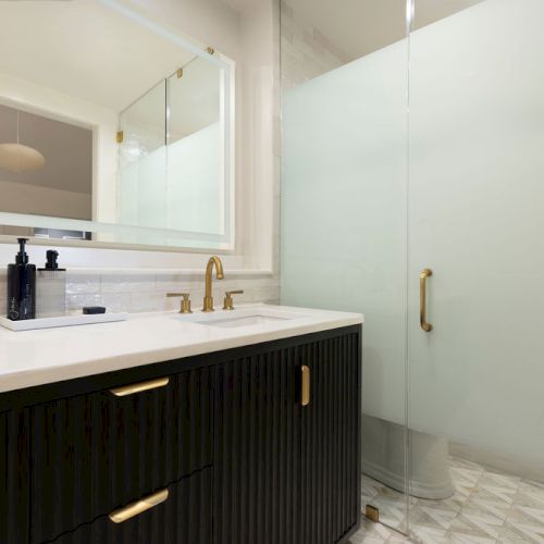 A modern bathroom with a black vanity, gold fixtures, a mirror, and a walk-in shower with a frosted glass door is shown.