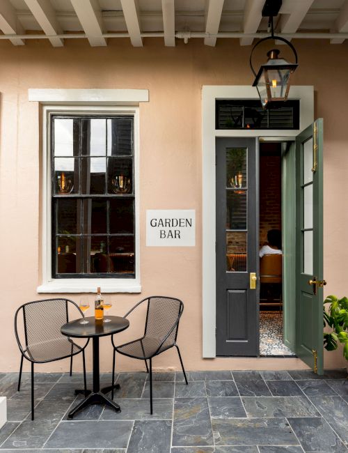 A small outdoor seating area with two chairs and a table, featuring a bottle, near a window and door labeled "Garden Bar."