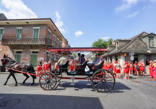 A horse-drawn carriage moves through a street with people dressed in red nearby and historic buildings in the background.