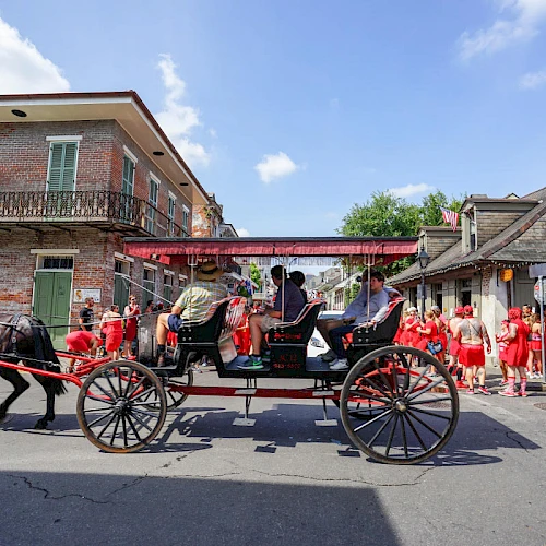 A horse-drawn carriage moves through a street with people dressed in red nearby and historic buildings in the background.