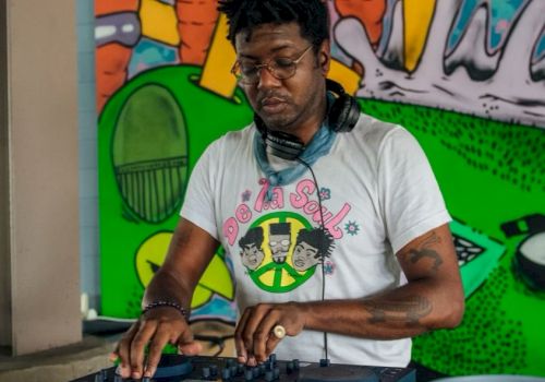 A person is DJing with headphones, in front of a colorful mural featuring cartoon carrots and other abstract elements.