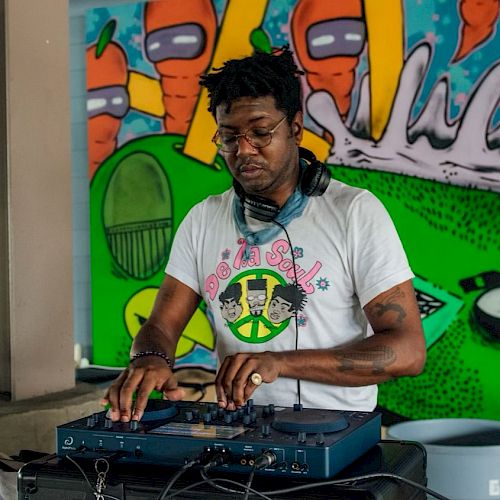 A person is DJing with headphones, in front of a colorful mural featuring cartoon carrots and other abstract elements.