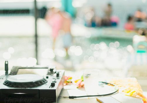 A turntable sits on a table by a pool, with blurred people in the background and sunny reflections on the water.