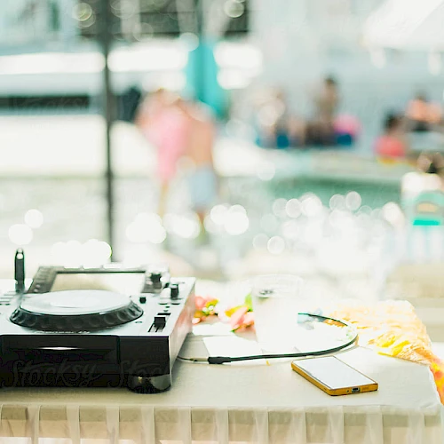 A turntable sits on a table by a pool, with blurred people in the background and sunny reflections on the water.