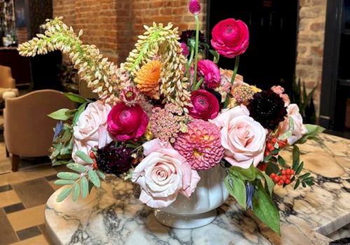 A vibrant flower arrangement in a white vase on a marble table, featuring pink roses, bright ranunculus, and greenery against a brick wall backdrop.