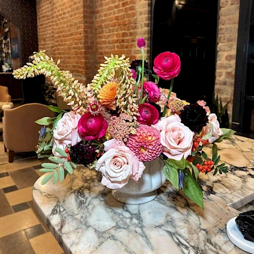 A vibrant flower arrangement in a white vase on a marble table, featuring pink roses, bright ranunculus, and greenery against a brick wall backdrop.