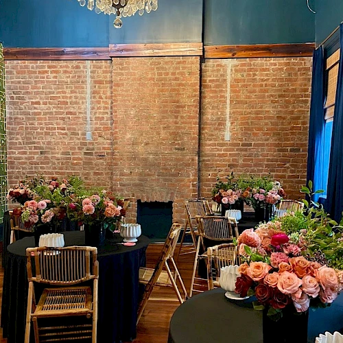 A brick-walled room with tables, flowers, and chairs, featuring a chandelier hanging above, creating an elegant atmosphere.