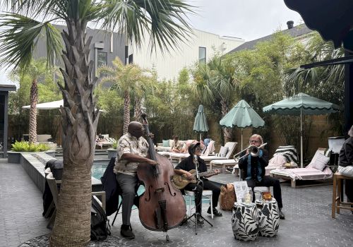 A musician plays a double bass near a poolside area with palm trees, lounge chairs, and string lights overhead, creating a relaxing atmosphere.