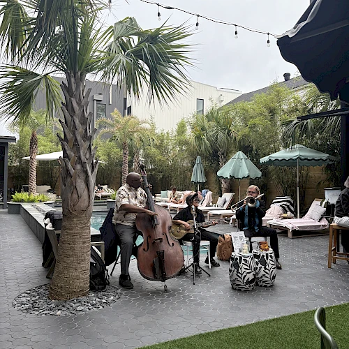 A musician plays a double bass near a poolside area with palm trees, lounge chairs, and string lights overhead, creating a relaxing atmosphere.