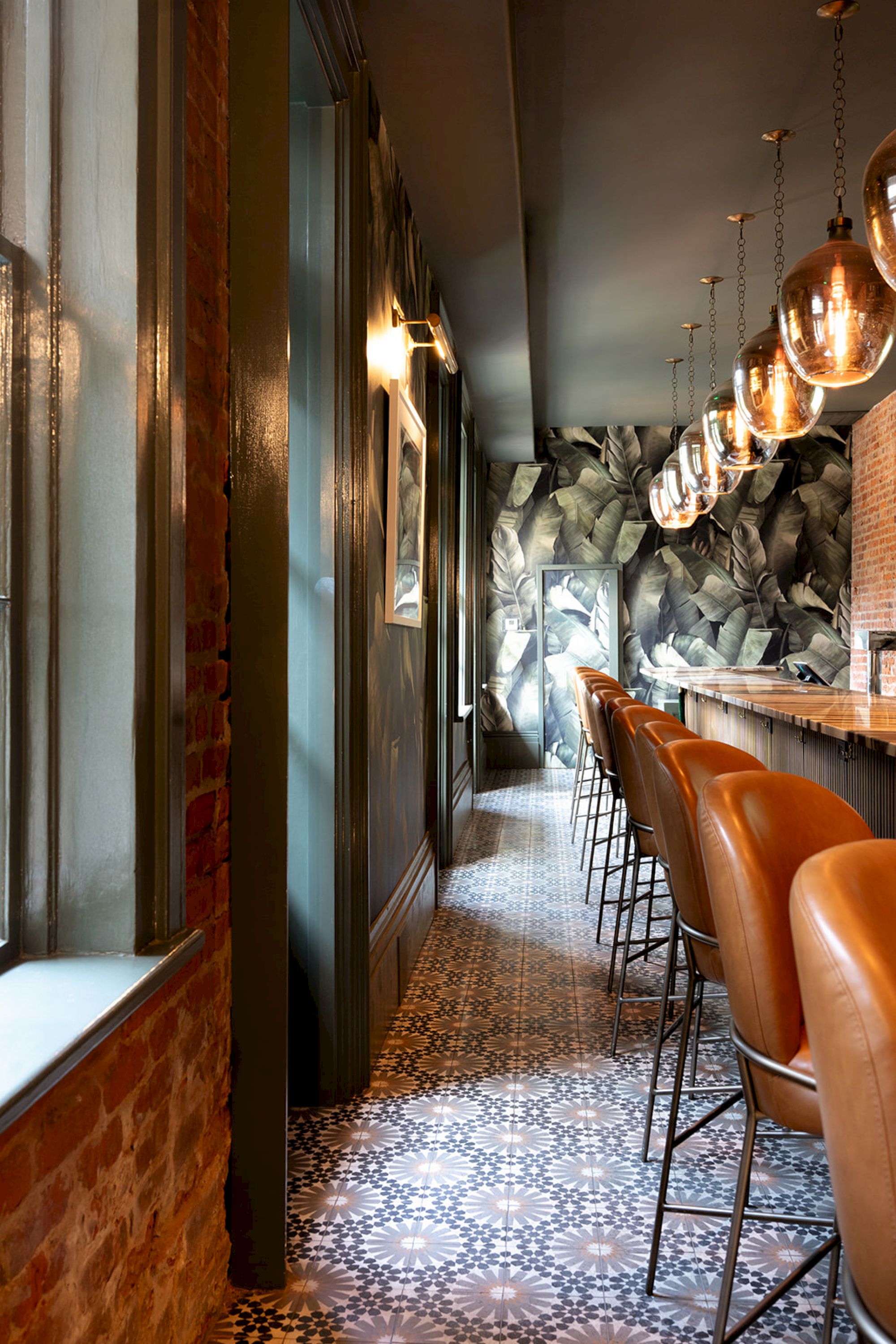 The image shows a stylish bar with leather chairs, pendant lights, and patterned floor tiles, featuring a wall with artistic decor.