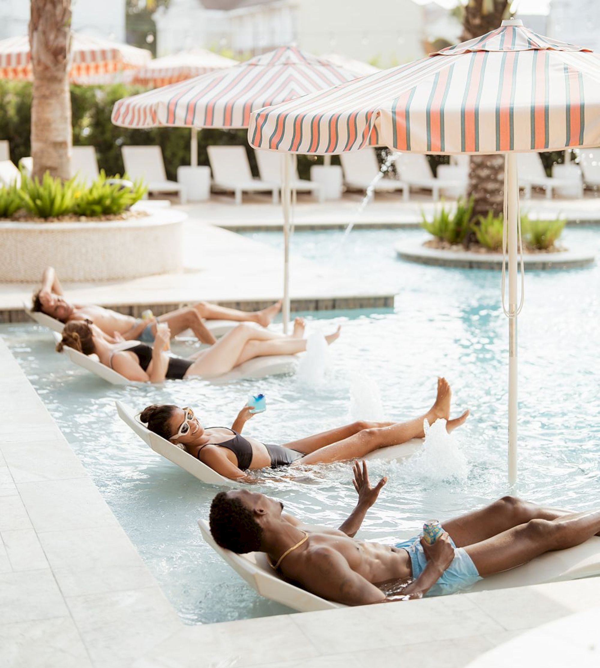 People are relaxing on loungers in a pool under striped umbrellas, enjoying a sunny day with drinks.