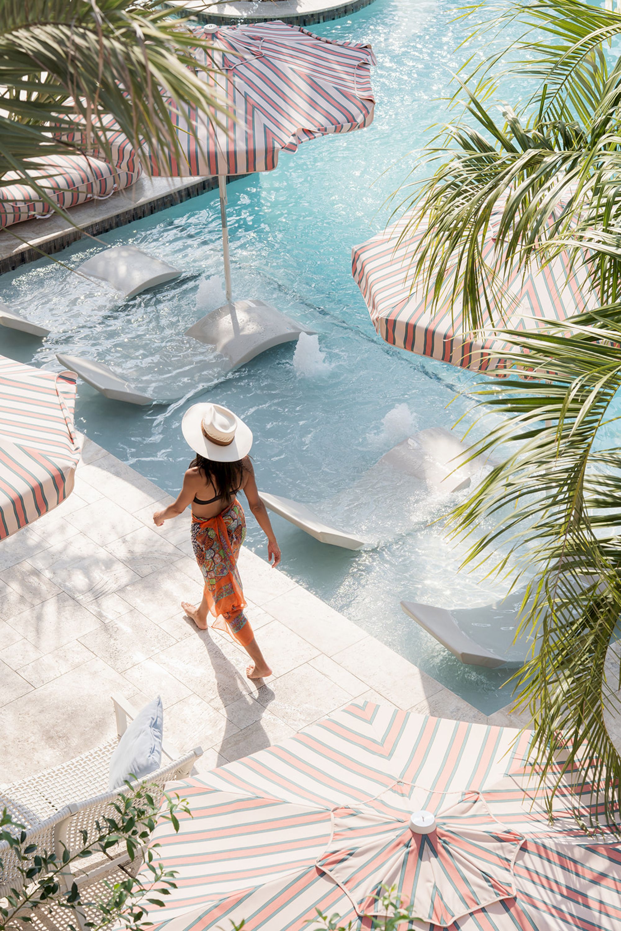 A person in a hat walks by a stylish pool with striped umbrellas, surrounded by palm trees, capturing a serene vacation vibe.