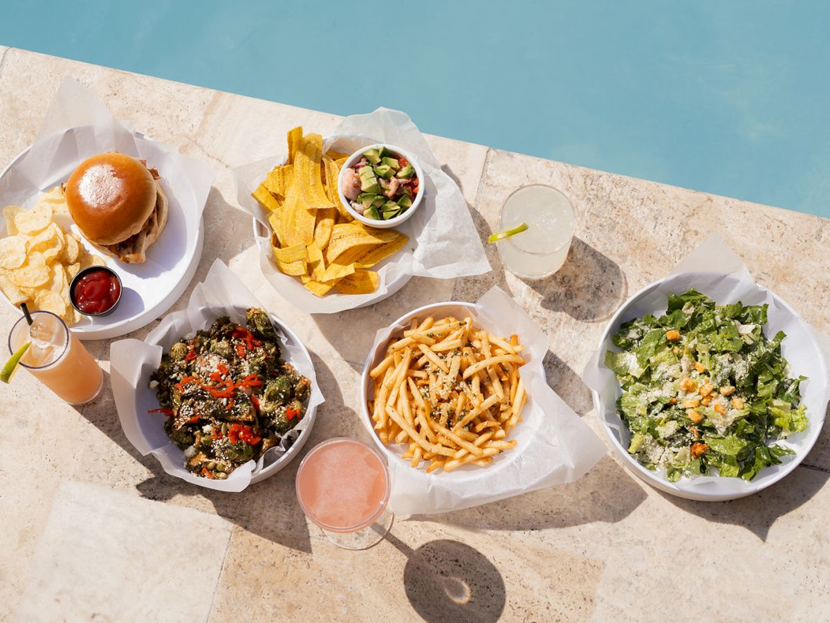 The image shows a variety of dishes including a burger, nachos, fries, a salad, and drinks, all set by a pool.