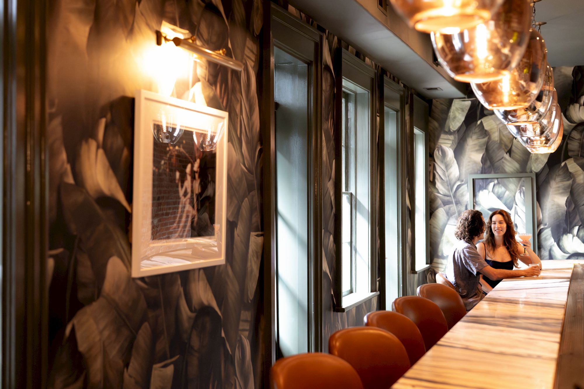 The image shows a dimly lit lounge with patterned walls, a long marble table, brown chairs, and two people sitting at the far end.