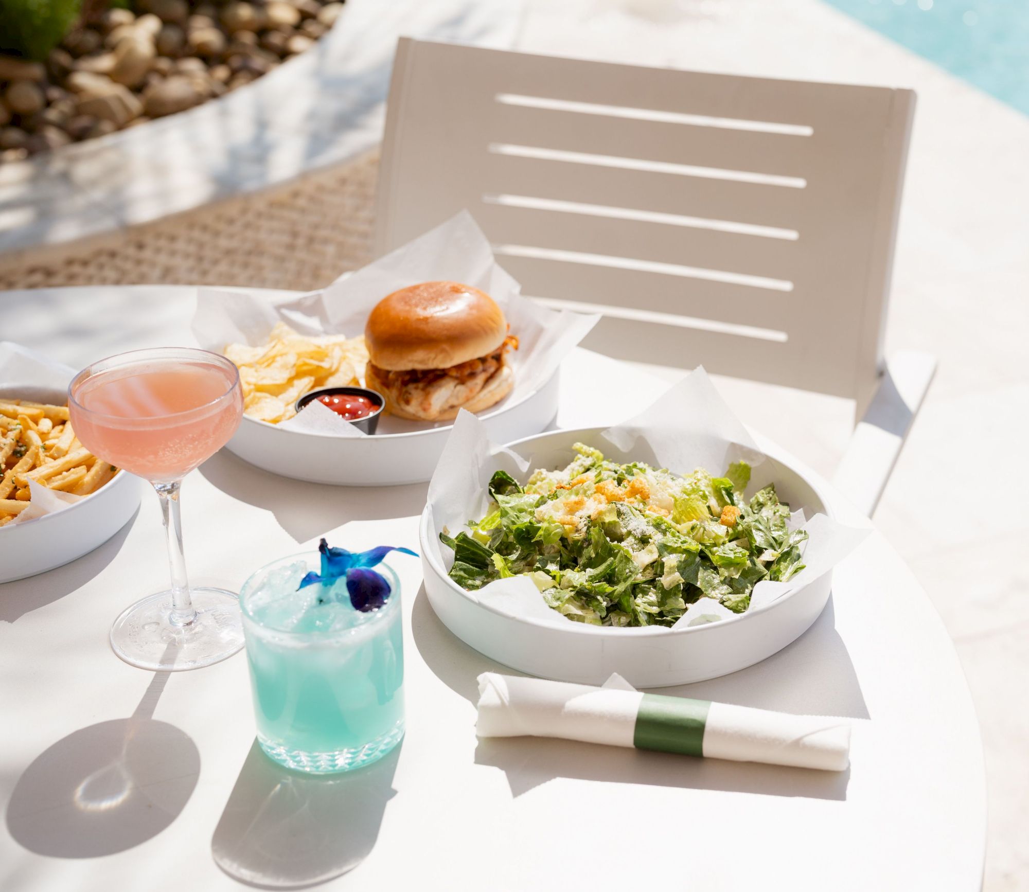 A table with a salad, burger, fries, and two colorful drinks by a poolside setting.