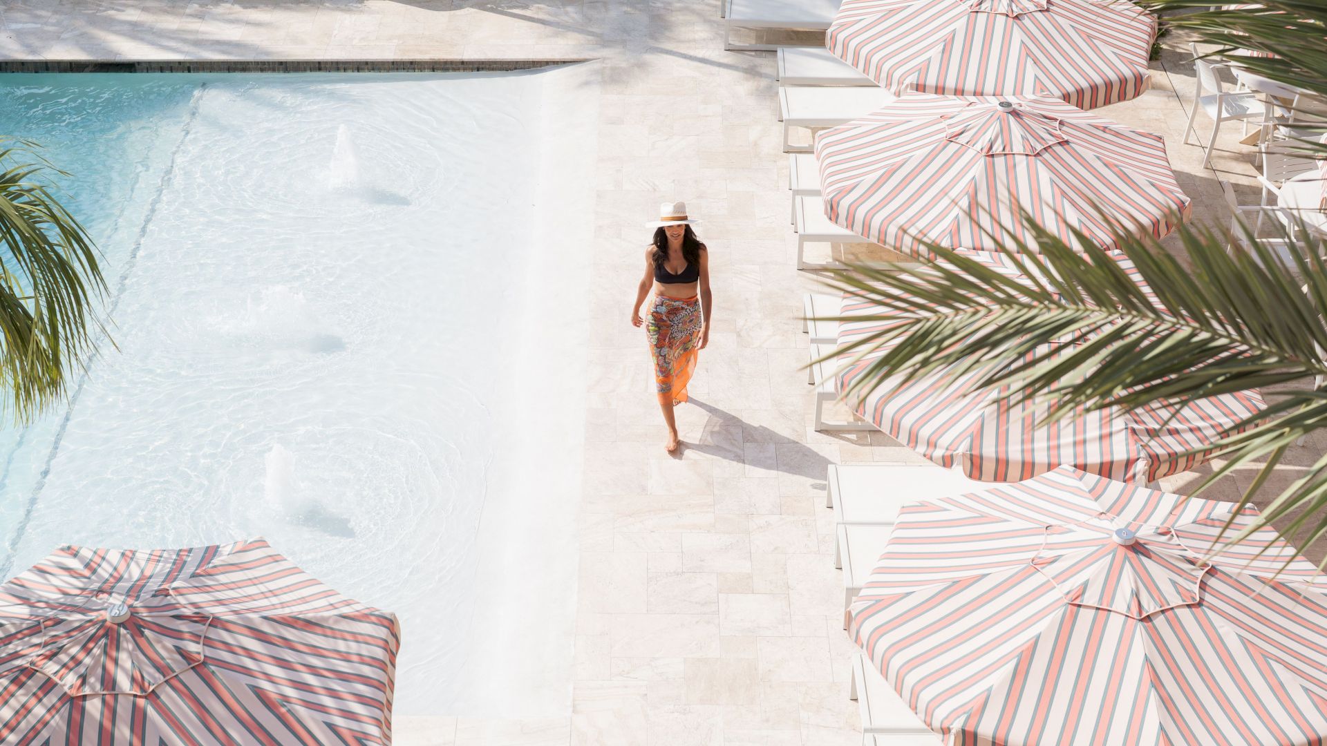 A person in a hat walks by a pool with striped umbrellas and palm leaves in a sunlit setting.