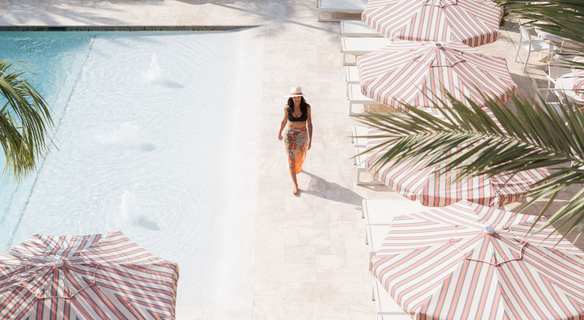 A person in a hat walks by a pool with striped umbrellas and palm leaves in a sunlit setting.