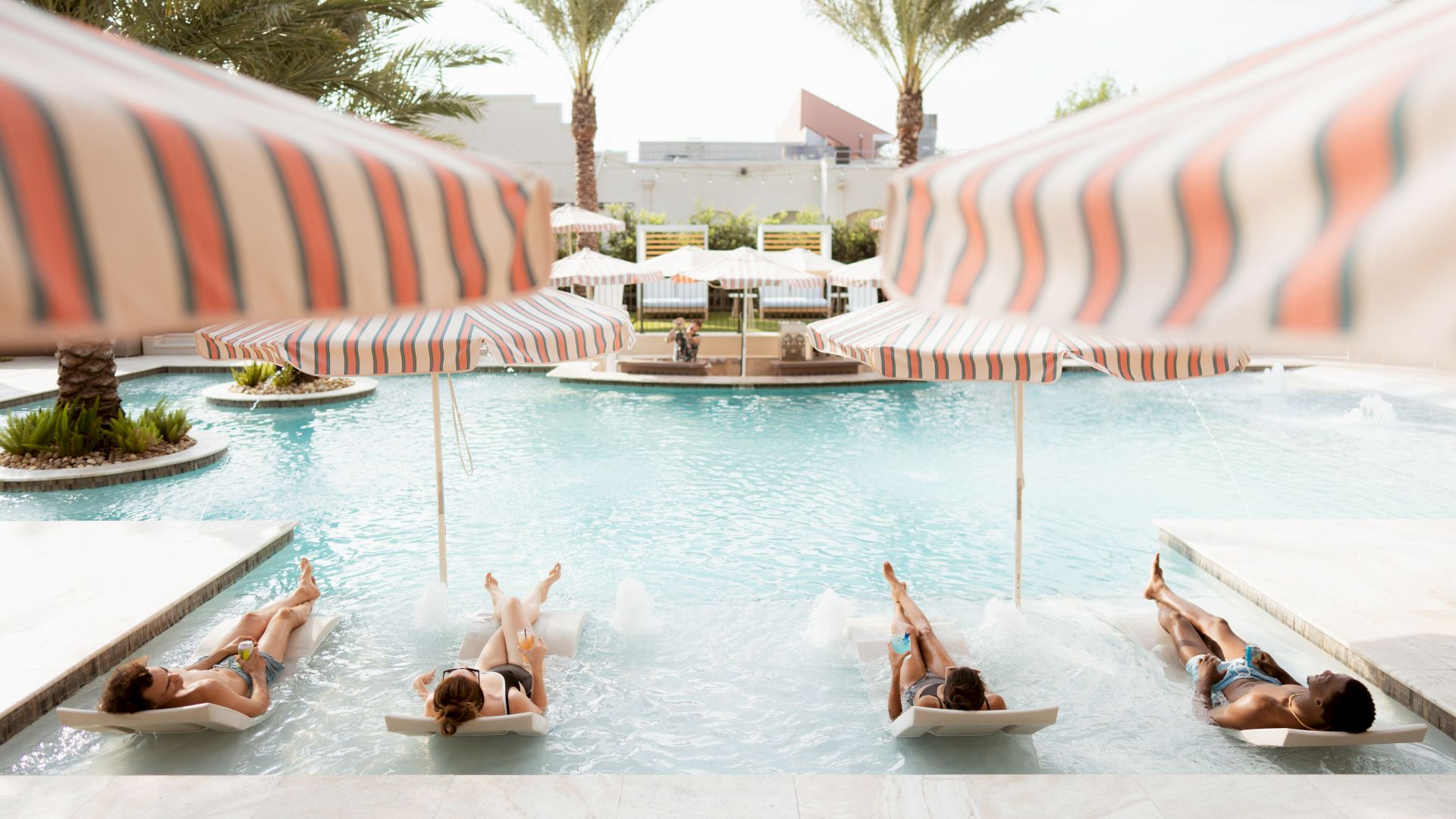 People are lounging on chairs in a pool under striped umbrellas, surrounded by palm trees and a serene resort setting.