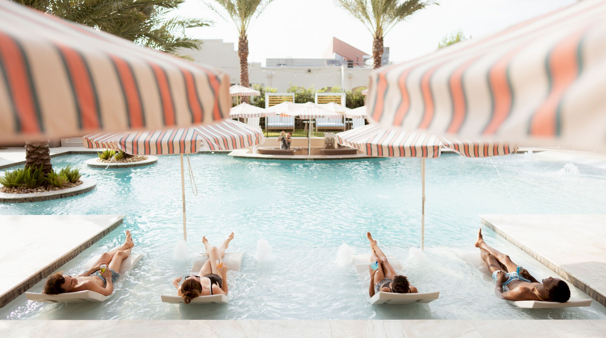 People are lounging on chairs in a pool under striped umbrellas, surrounded by palm trees and a serene resort setting.