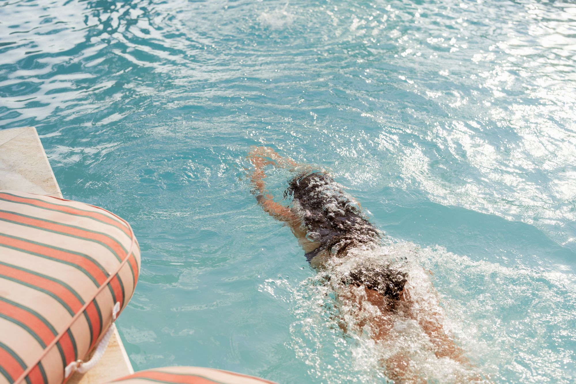 A person is swimming in a pool near the edge with striped cushions visible, creating splashes as they move through the water.