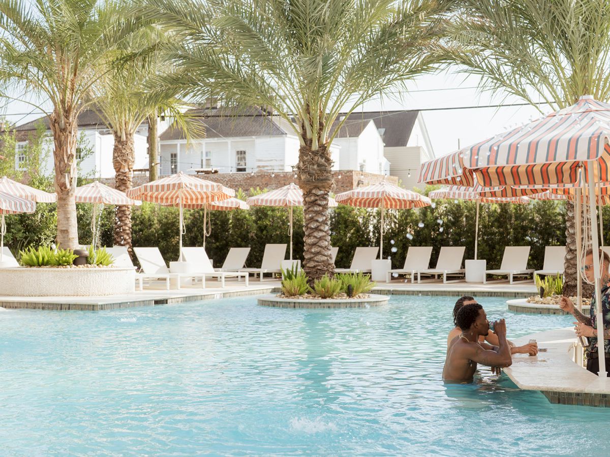 A sunny pool area with palm trees and striped umbrellas, with people relaxing in the water and on lounge chairs.
