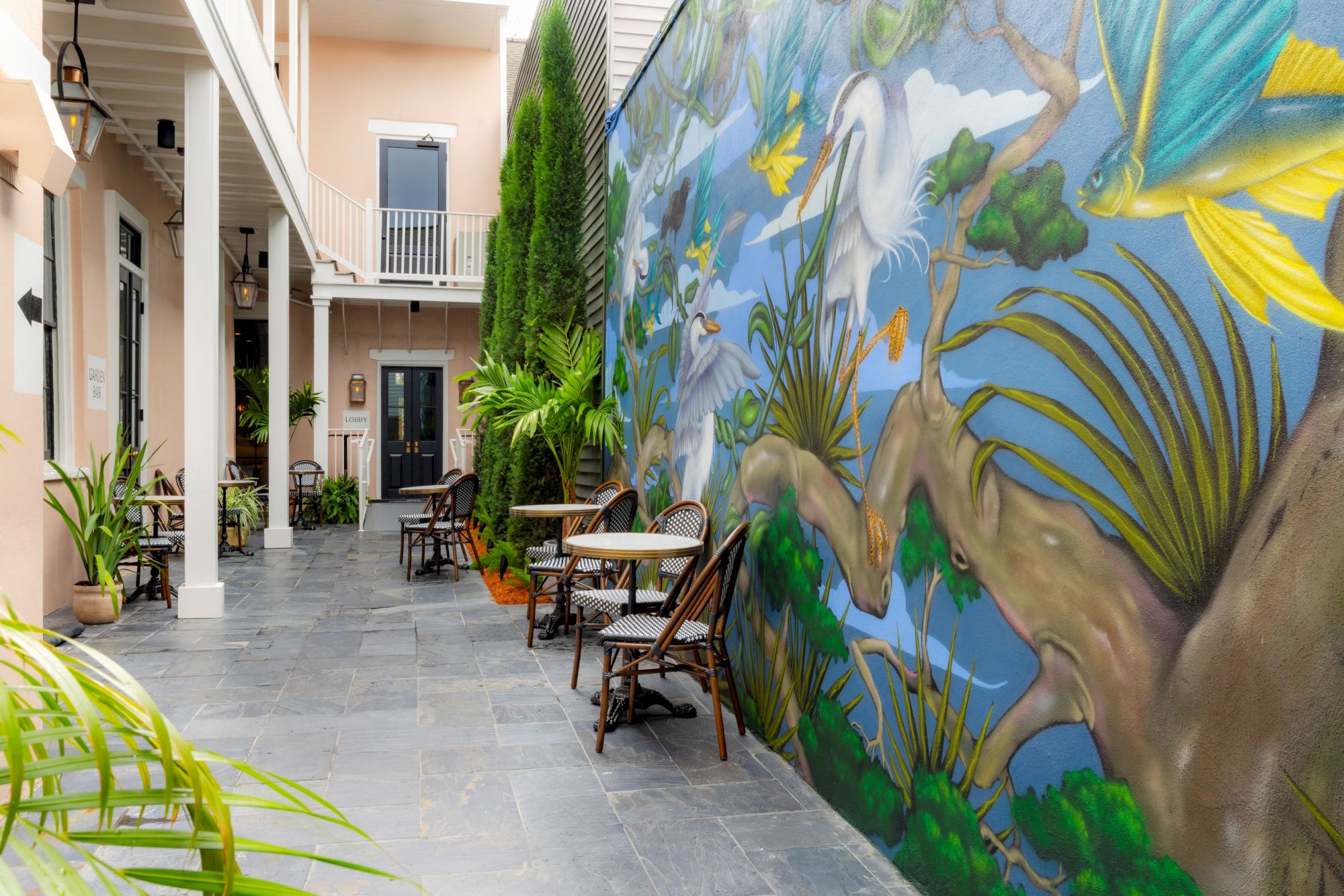 An outdoor patio with tables and chairs, lined by a vivid mural of birds and nature on the wall, next to a building.