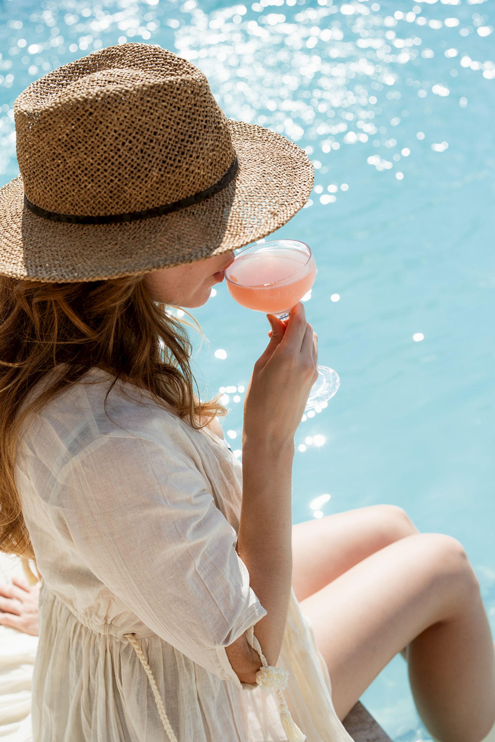 A person wearing a straw hat and light clothing sits by a pool, sipping a pink cocktail.