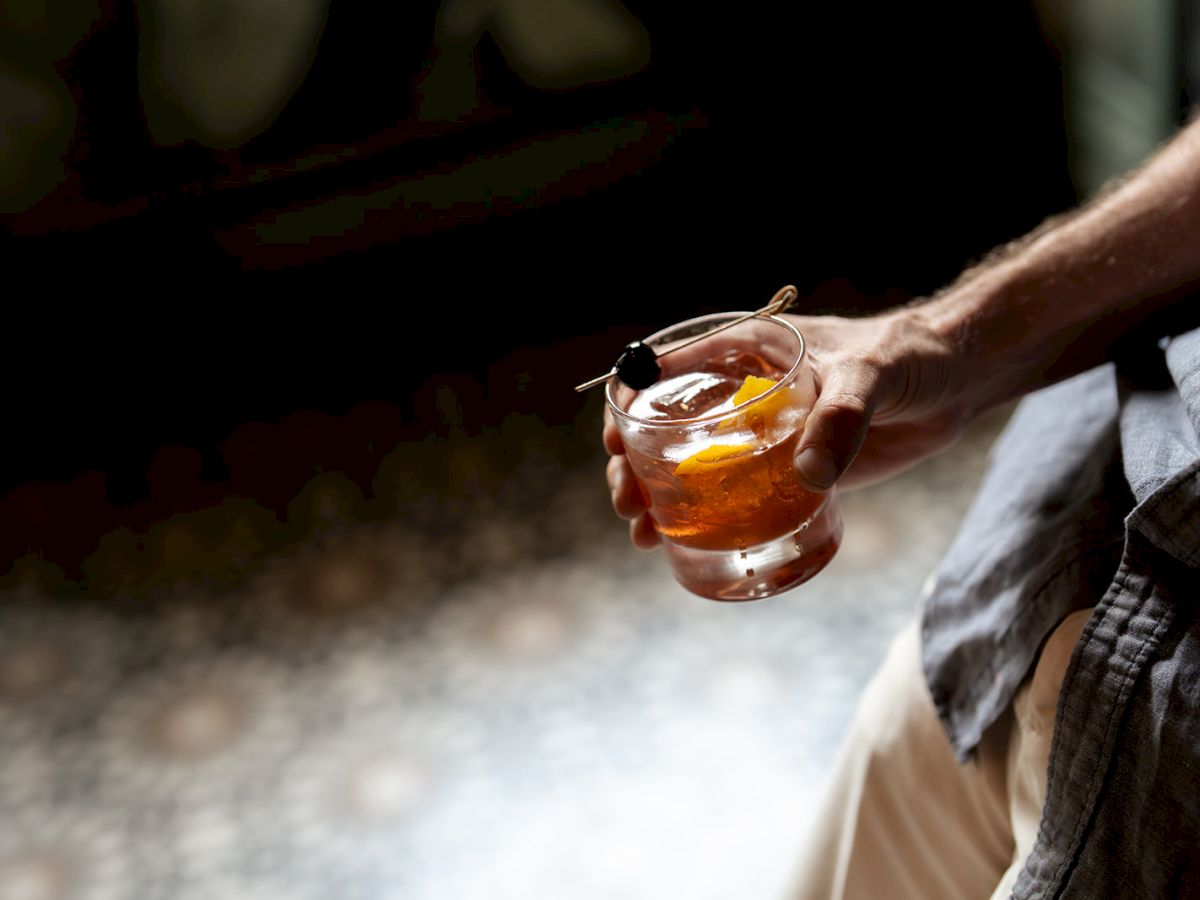 A person is holding a glass of dark amber whiskey or whiskey-like drink with ice, outdoors, near a cobblestone surface, in partial shade.