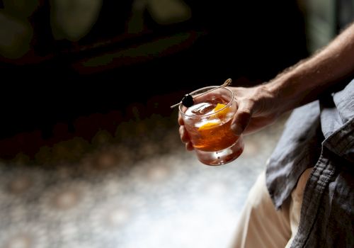 A person holding a short glass with whiskey or whiskey-like drink and an ice cube, outdoors, hand visible, casual attire, relaxed vibe.
