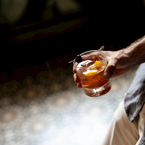 A person holding a short glass with whiskey or whiskey-like drink and an ice cube, outdoors, hand visible, casual attire, relaxed vibe.