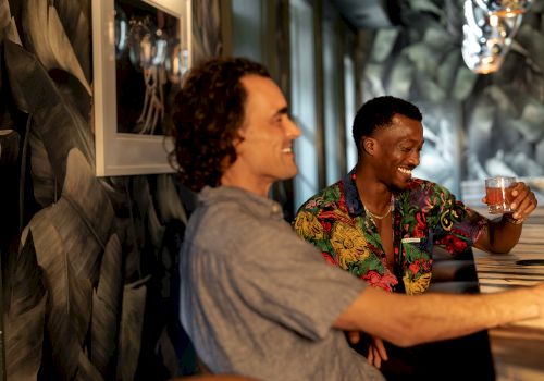 Two friends laugh in a cozy bar, one holding a drink in a colorful shirt while the other smiles across the table.