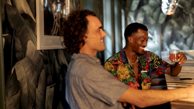 Two friends laugh in a cozy bar, one holding a drink in a colorful shirt while the other smiles across the table.