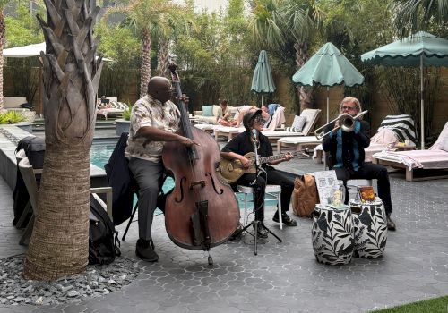 A trio of musicians playing outdoors by poolside: a bassist, a guitarist, and a saxophonist or vocalist, under palm trees and umbrellas.