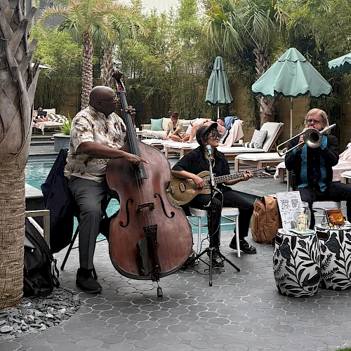 A trio of musicians playing outdoors by poolside: a bassist, a guitarist, and a saxophonist or vocalist, under palm trees and umbrellas.