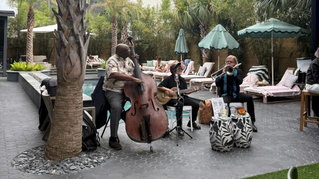 A trio of musicians playing outdoors by poolside: a bassist, a guitarist, and a saxophonist or vocalist, under palm trees and umbrellas.